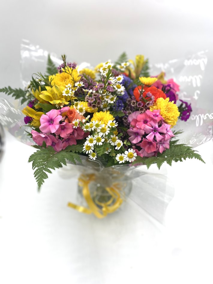 זר פרחים צבעוני עם פרחים ורודים, צהובים, כתומים, לבנים וסגולים, מקושט בעלי שרך ירוקים ועטוף בניילון שקוף עם סרט צהוב.