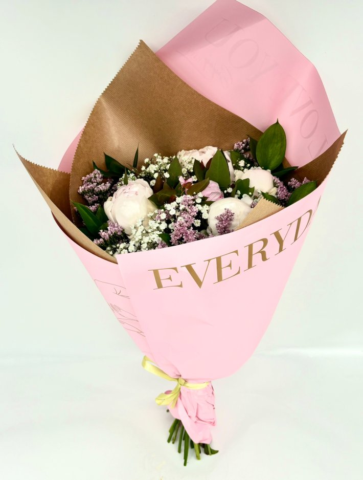 זר פרחים עדין עם פרחי פיאונים לבנים, פרחים סגולים קטנים ועלים ירוקים, עטוף בנייר קראפט חום ונייר ורוד עם כיתוב אלגנטי