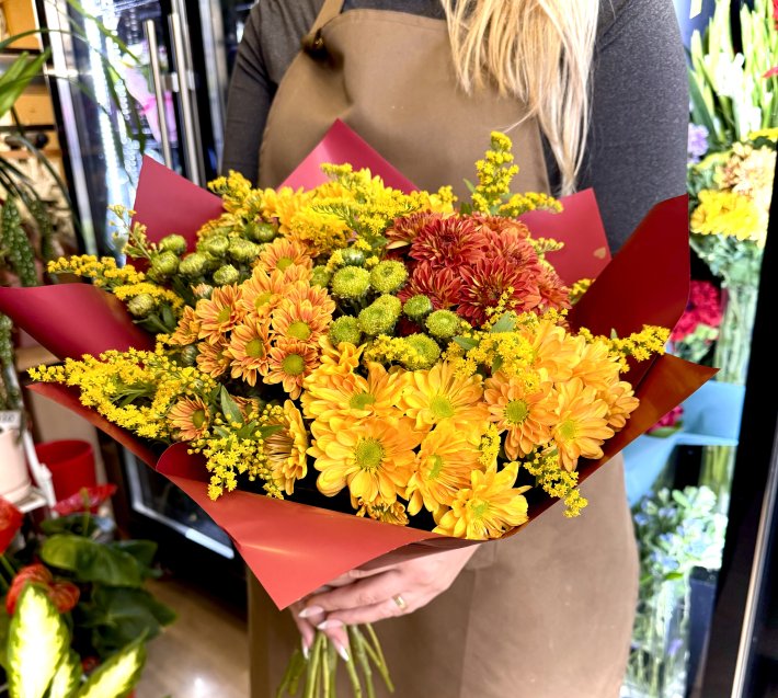 זר פרחים סתווי הכולל פרחים כתומים, צהובים ואדומים עם תוספות ירוקות, עטוף בנייר אדום מהודר, מושלם לאווירה חמה וחגיגית